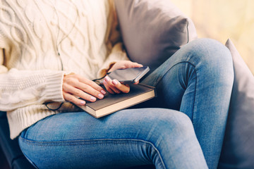 Young girl in a white sweater and jeans sitting on the window sill holding a smartphone in hands and listening to music