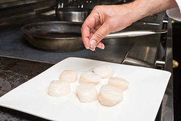 Delicious  organic scallops on a white plate, being hand seasoned with sea rock salt, in preparation for pan cooking.