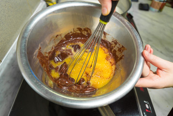 A metal bowl on a hob, with butter and dark chocolate cooking peices melting into a sauce, in a metal industrial kitchen.