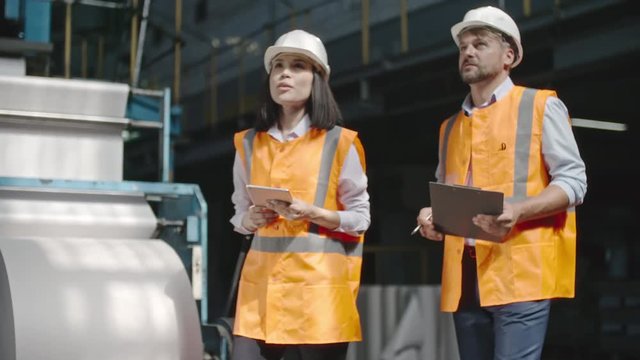 Female chief engineer in reflective safety vest inspecting factory workshop and discussing it with male bearded colleague