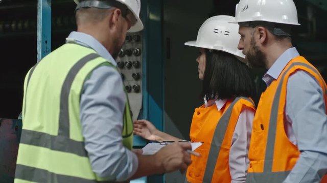 Female engineer in hard hat and safety vest standing before industrial machine and describing process of its work to male colleague with clipboard making notes 