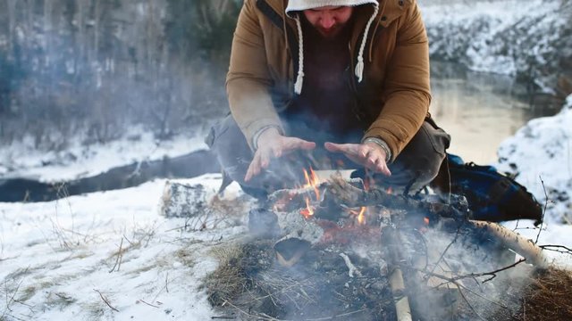 Tilt down of man sitting by fire on camping trip in winter rubbing his cold hands together and trying to warm up