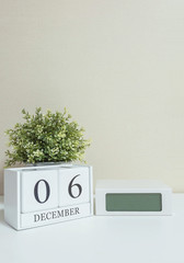 White wooden calendar with black 6 december word with clock and plant on white wood desk and cream wallpaper textured background , selective focus at the calendar