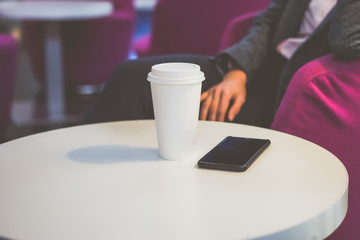 Close-up cup of coffee and a smartphone on a white round table. The photo shows a part of the girl in a jacket, sitting in a coffee shop on a pink chair next to the table. Blurred background.