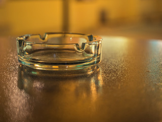 Picture of the glassy ashtray close up. Ashtray laying on the blurred surface of the table. Blurred background of the ashtray on the table. Blurred surface of the table reflects yellow sunlight.