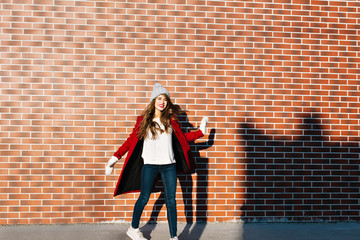 Fototapeta na wymiar Beautiful brunette girl with long hair in red coat and knitted hat on wall background outside. She is turning round in sunshine and smiling to camera
