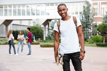 Cheerful african man student with backpack holding skateboard outdoors