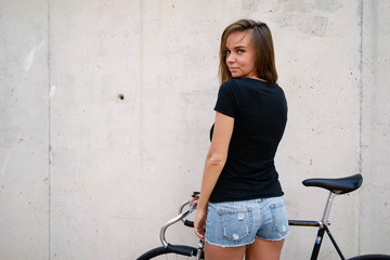 A brunette girl with long hair wearing a blank black t-shirt is standing with the back to the camera on a gray wall background on a street. Empty space for text or design. - 128617237