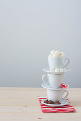 A stack of cups and saucers with hot chocolate ingredients: chocolate, mini marshmallow, peppermint candy cane. 