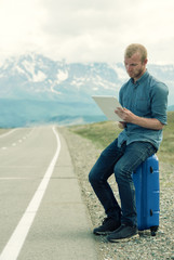 Alone man sits on a suitcase and looking at tablet. Waiting for  bus or  car. The road in the mountains.