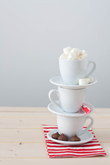 A stack of cups and saucers with hot chocolate ingredients: chocolate, mini marshmallow, peppermint candy cane. 