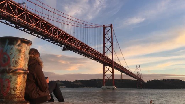 Girl With Smartphone Sitting By Tagus River Enjoying Sunset, Lisbon. The 25 de Abril Bridge is a suspension bridge connecting the city of Lisbon to the municipality of Almada.