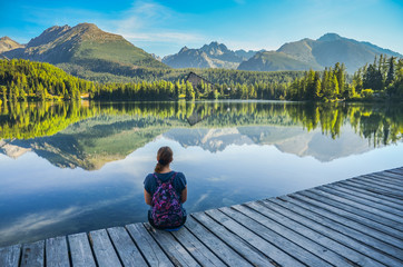 Alone young girl sitting and resting on the wooden path near by beautiful blue lake and clear big mountains. Original wallpaper from summer morning
