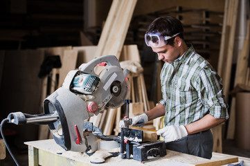 Skilled carpenter cutting a piece of wood in his woodwork workshop, using circular saw with other machinery on the background