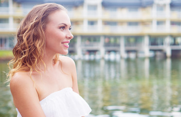 Side closeup face of pretty blond woman in white summer dress standing on the pier and smiling against blurred lake and tropical resort background. Beautiful female enjoying her holiday