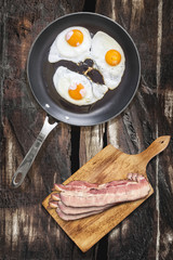 Sunny Side Up Fried Eggs In Heavy Duty Teflon Frying Pan With Pork Bacon Rashers On Cutting Board Set On Old Cracked Wooden Picnic Table