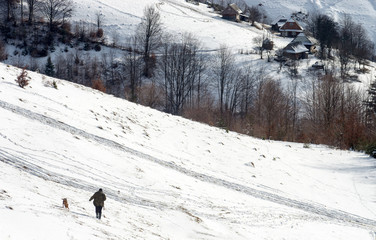Hunter with a dog and a shotgun walking on snow-covered hill to the wooden cabins. Winter hunting.