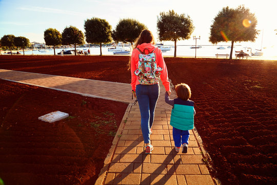 Young woman and little toddler boy walking through a park to the sea. Mother in orange jacket with backpack and small son go to the quay with fish boats and yachts. Beautiful green trees on sunset.