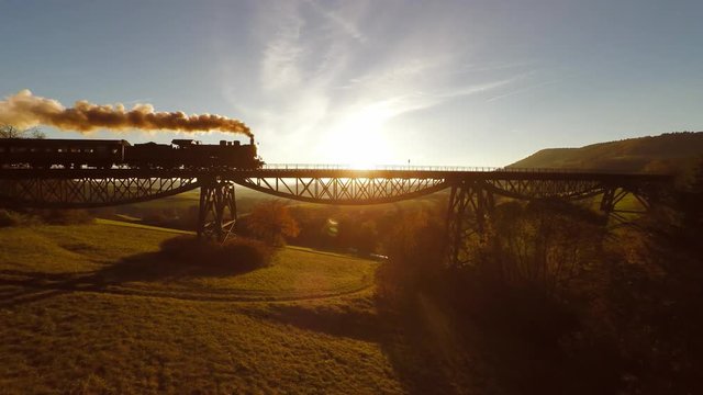 aerial view of steam engine locomotive crossing bridge at sunset magic hour. Epic scenery background.