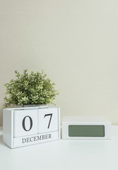 White wooden calendar with black 7 december word with clock and plant on white wood desk and cream wallpaper textured background , selective focus at the calendar