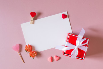 Valentine day composition: red gift box with bow, stationery / photo / postcard template with clamp, small hearts, candy, and spring flowers on light pink background. Top view.