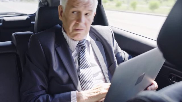 Senior businessman in moving car checking his wristwatch and working on laptop