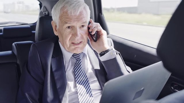 Grey haired businessman working on laptop and talking with someone over mobile phone in moving car
