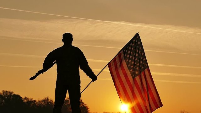 
Soldier lift up rifle and hold American flag against  orange sky. Slow Motion close up
