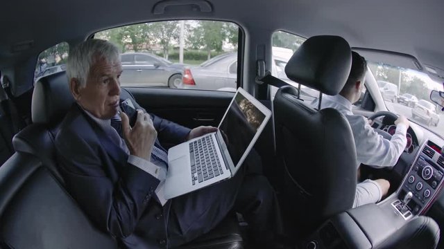 Fish eye shot of senior businessman sitting in moving car working on laptop and talking on mobile phone with speakerphone 