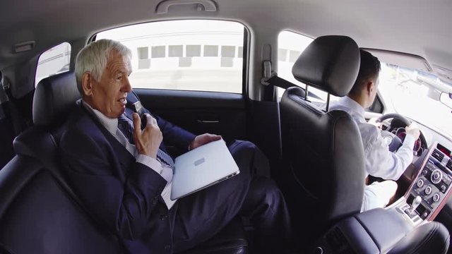 Fish eye shot of male senior businessman sitting in car with personal chauffer and talking on mobile phone via speakerphone