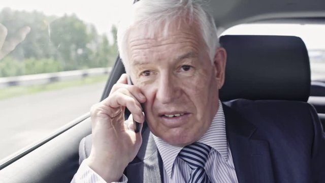 Senior businessman sitting in moving car and talking on mobile phone
