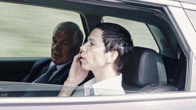 Shot from another vehicle of moving car with businesswoman talking on mobile phone and sitting next to senior colleague with tablet