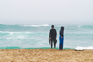 Two surfers stand ashore ocean. Fellow and girl.