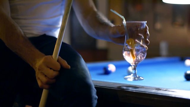 Man drinking alcoholic drink while sitting on billiard's table and holding cue
