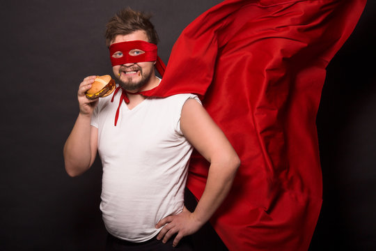 Super hero glutton man eating hanburger isolated on black. Handsome man with red mask on posing for photographer. Super hero concept.