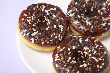 A plate full of iced chocolate sprinkle ring donuts on a pastel purple background