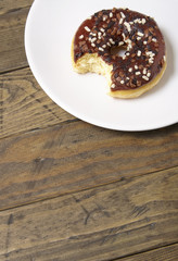 An iced chocolate sprinkle ring donut with bite mark, on a rustic wooden background with blank space below