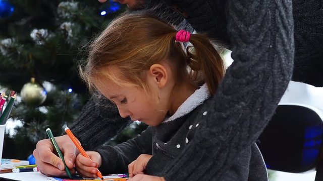 Father helping her daughter in drawing. Watching little girl drawing on paper. Christmas lights background