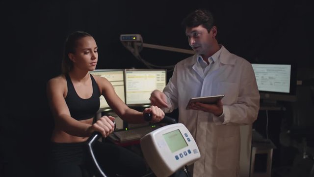 Doctor in lab coat holding tablet and explaining something to sporty woman sitting on exercise bike