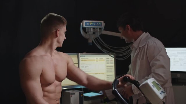 Tilt up of handsome shirtless man with muscles sitting on exercise bike and chatting with doctor placing electrodes on his body