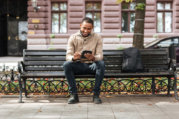Man with tablet sitting on bench near backpack and typing