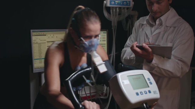 Doctor writing down results of stress testing of exhausted female athlete on exercise bike