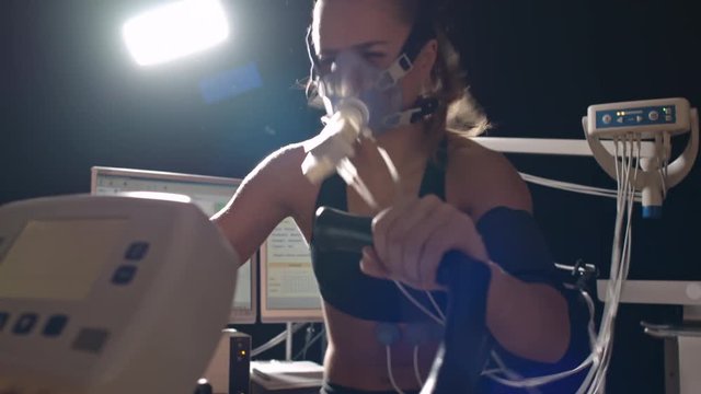 Tilt up of strong woman in oxygen mask cycling exercise bike in darkly lit laboratory