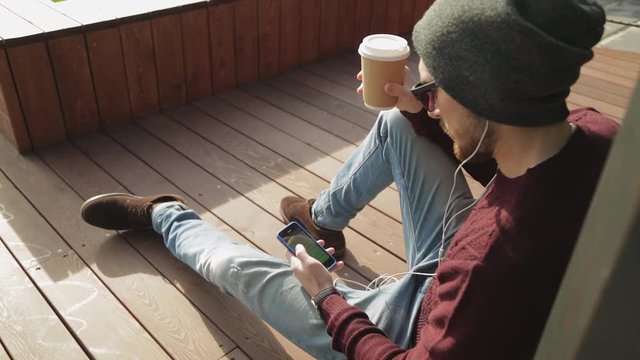 Handsome hipster listening to music in smartphone headphones and drinking coffee from paper сup