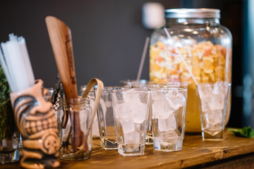 Several glasses with ice cubes, a jar of orange slices and citrus juice on the wooden bar