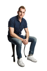 A sad man sitting down on a stool in front of a white background, wearing a blue shirt and jeans with white shoes, looking at camera.