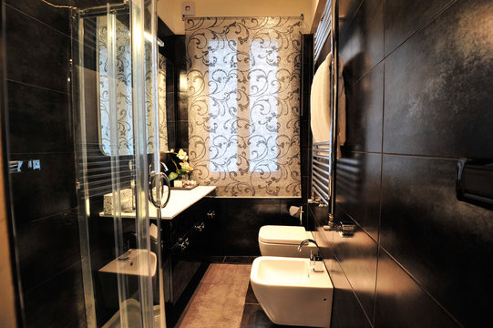 Italian Bathroom With Black Wall / Tiles And White Forniture / Services. A Warm Light Illuminates The Room