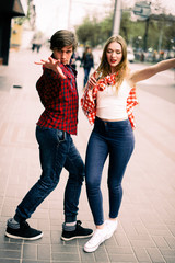 Fototapeta premium Two happy trendy teenage friends walking and dancing in the city, listening to the music with headphones, talking each other and smiling. Lifestyle, friendship and urban life concepts.