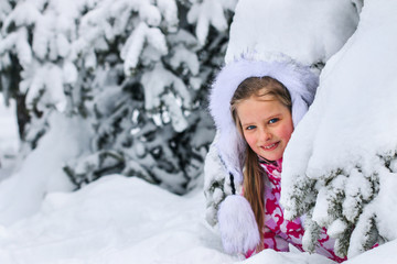 Portrait of little kid girl in winter clothes plying in deep snow uder fir tree.