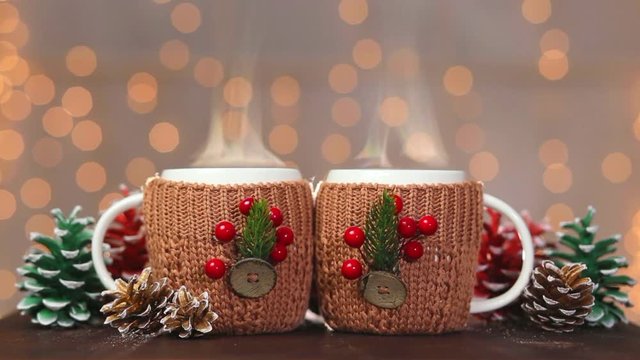 Close up of two white steaming cups in handmade brown cute knitted covers with festive christmas decorations at blurry lights of orange garland background. Real time full hd video footage.